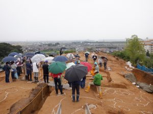 観法寺墳墓群でみつかった弥生時代後期の竪穴建物について、現地説明会で小雨の中、多くの見学者が説明をきいているところです。